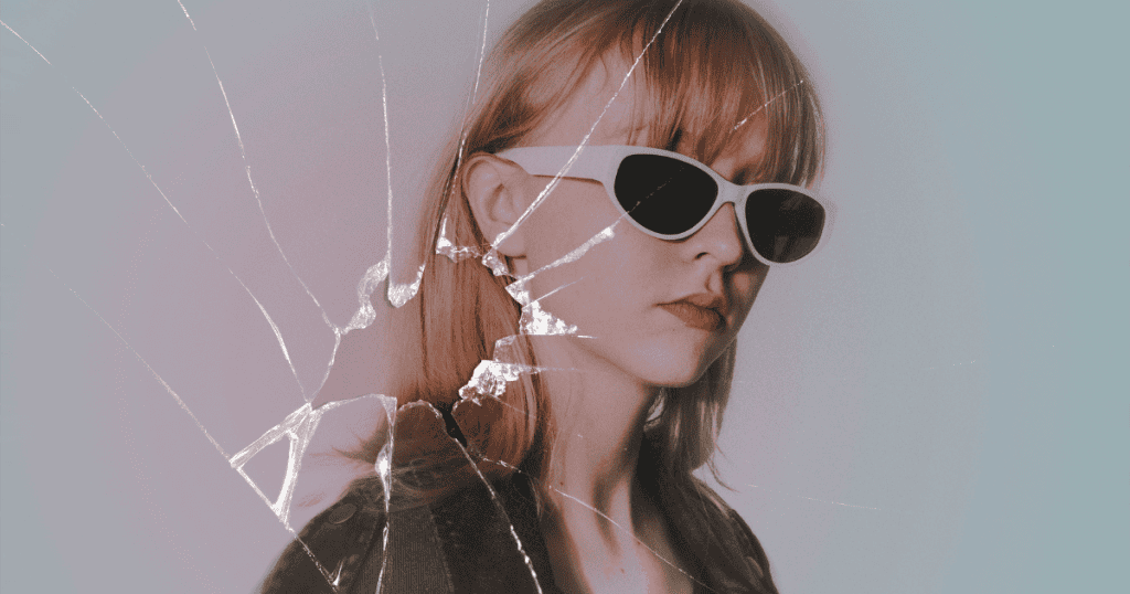 A woman with straight blond hair wearing white sunglasses and a black jacket is seen through cracked glass, symbolizing a fractured or trapped perspective. The cracks radiating across the image create a visual metaphor for breaking free from cycles of stress or distorted thinking.