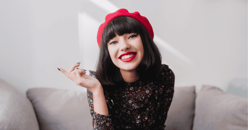 A smiling woman in a red beret and floral dress sits on a couch with confidence, symbolizing how to protect your starlight around narcissists.