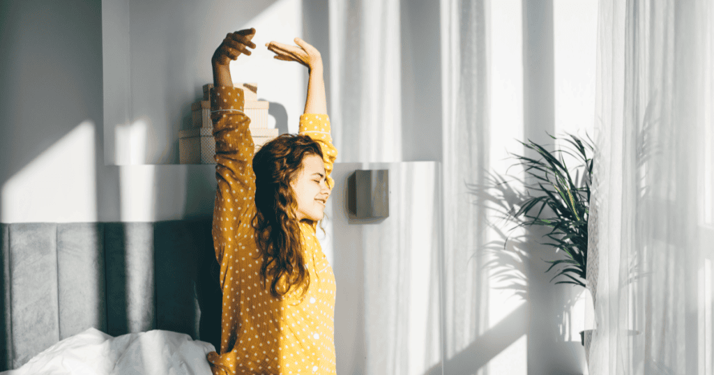 A woman stretches in bed with sunlight streaming through the window, taking rest without needing permission.
