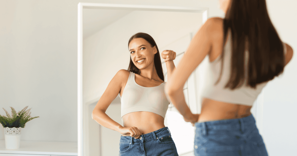 A smiling woman looks at herself in the mirror while wearing jeans and a crop top, celebrating her body after cutting ties with narcissists.