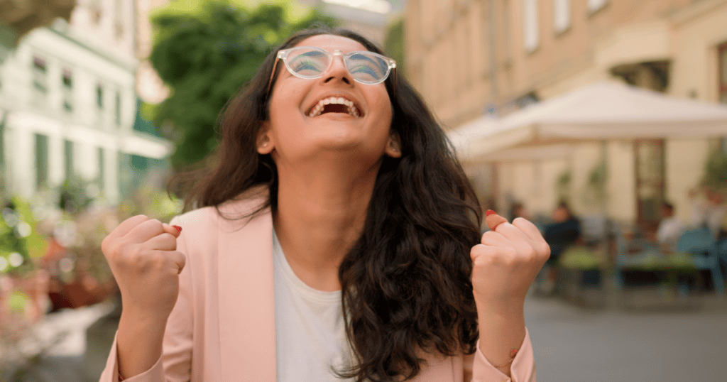 A joyful woman with glasses smiles and raises her fists in celebration, reclaiming her joy after cutting narcissists loose.