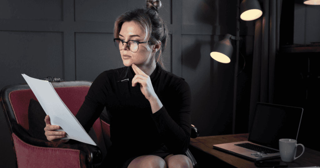 A woman in glasses studies a document thoughtfully while sitting in a dimly lit office, symbolizing the ability to see patterns before they play out.