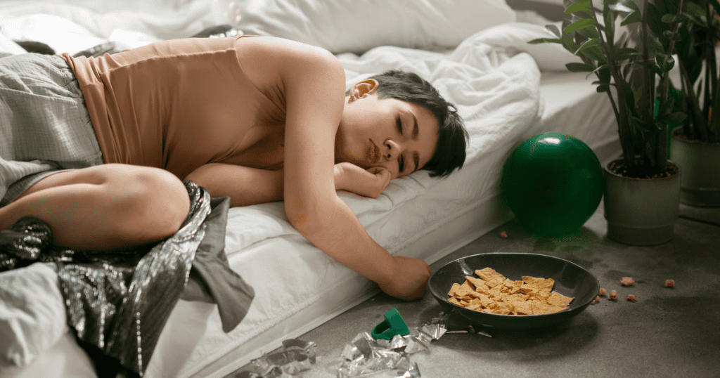 A person lies curled on a bed beside scattered snacks and empty wrappers, looking drained and disengaged, suggesting a tactic to deflate a narcissist’s adrenaline-fueled drama.