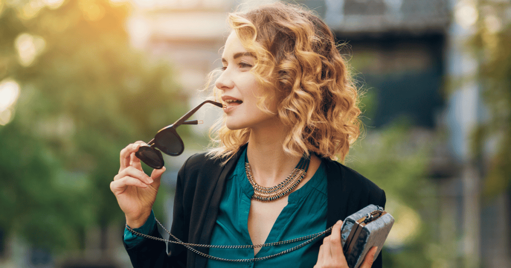 Confident woman with curly blonde hair holds sunglasses and smiles while walking outdoors in the sunlight, symbolizing empowerment and the strength that comes from embracing truth.