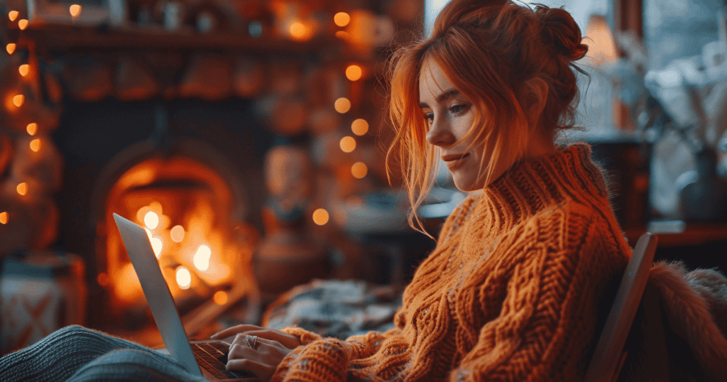 A woman in a cozy orange sweater working on her laptop by the fireplace, surrounded by warm lights, symbolizing personal truth and comfort instead of holding onto the false narratives of others.