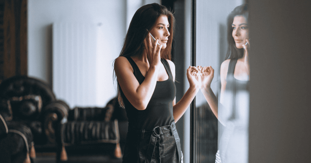 A woman stands by a window talking on her phone with her reflection visible in the glass; this portrays calm confidence and the idea that your comeback is the best revenge.