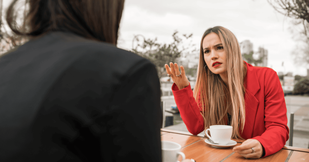 A woman in a red jacket gestures intensely while talking at a café, illustrating how narcissists gauge your listening style and reactions to control conversations.