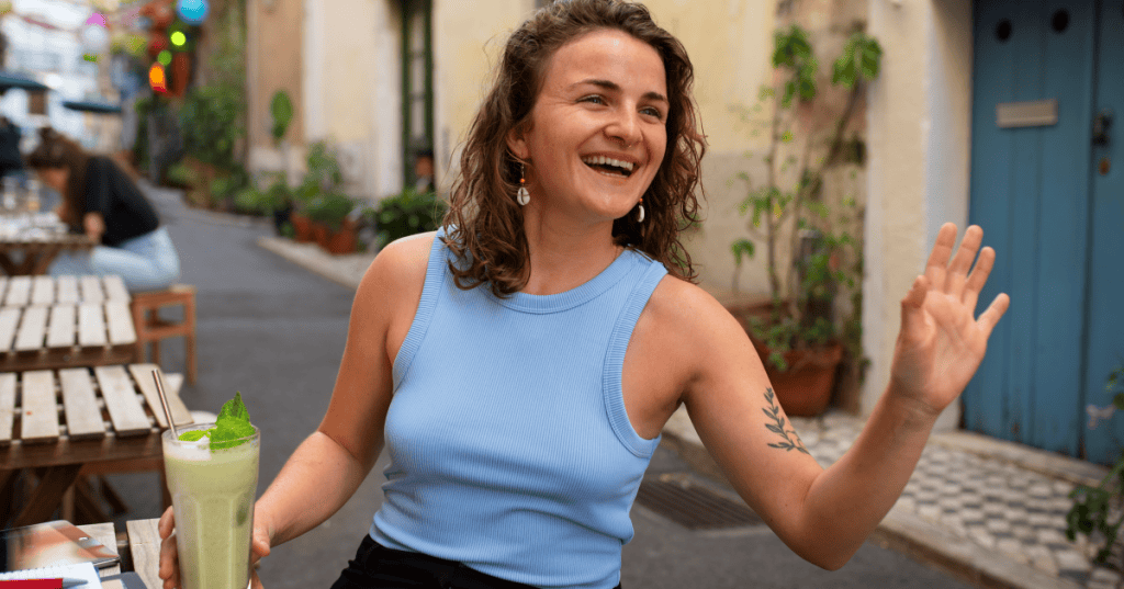 A woman waves goodbye with a smile while holding a drink, stepping away from the narcissist’s conversation.
