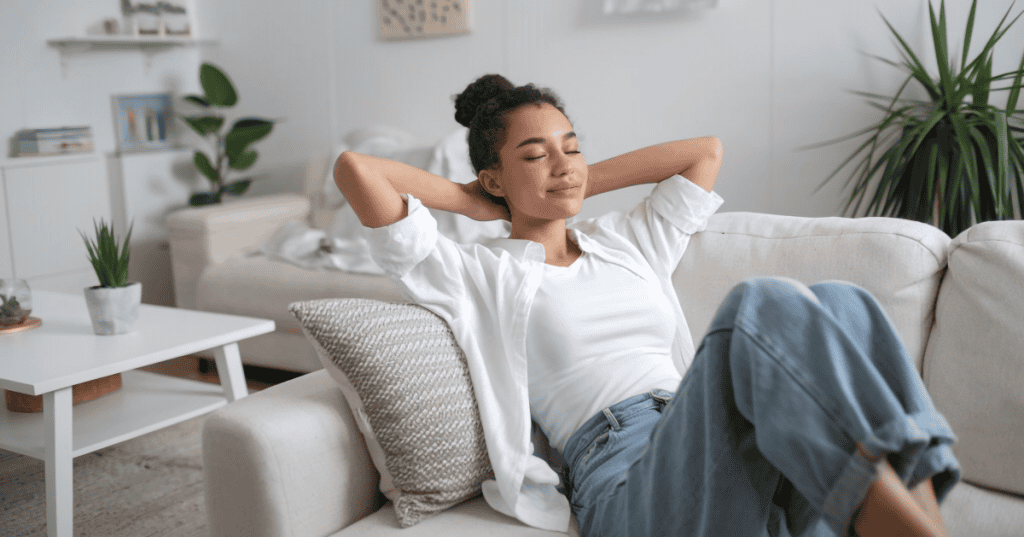 A relaxed woman leans back on a sofa with her eyes closed, resting without feeling the need to earn it.