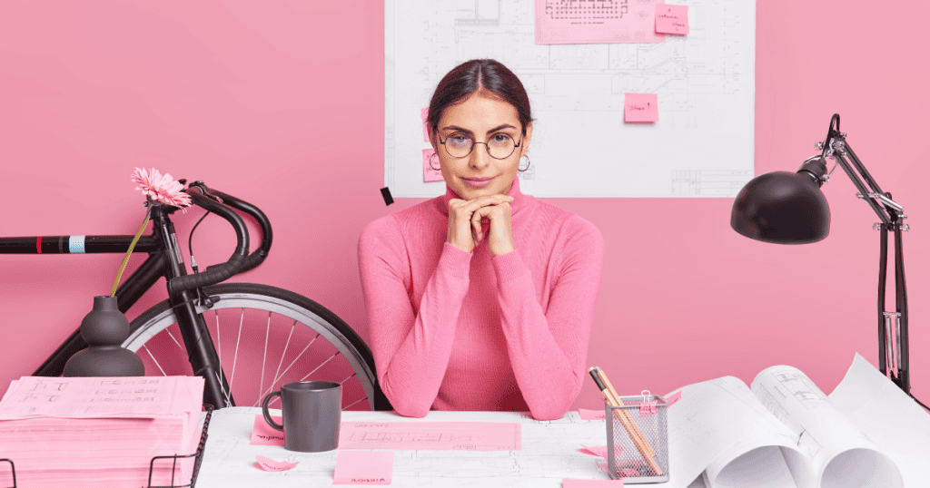 A woman in glasses sits confidently at a pink desk with plans and notes around her, symbolizing how narcissistic bosses rise in workplaces and why strategy is needed to outsmart them.