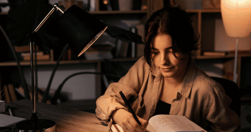 A young woman sits at a desk writing in a notebook under a lamp’s warm light, representing daily habits that keep you sharp and strong after narcissistic abuse.