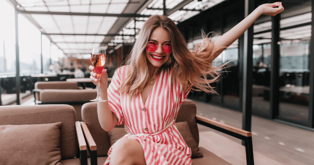 A smiling woman in a striped dress and sunglasses raises a glass while celebrating outdoors, capturing the confidence needed to say no to a narcissist.