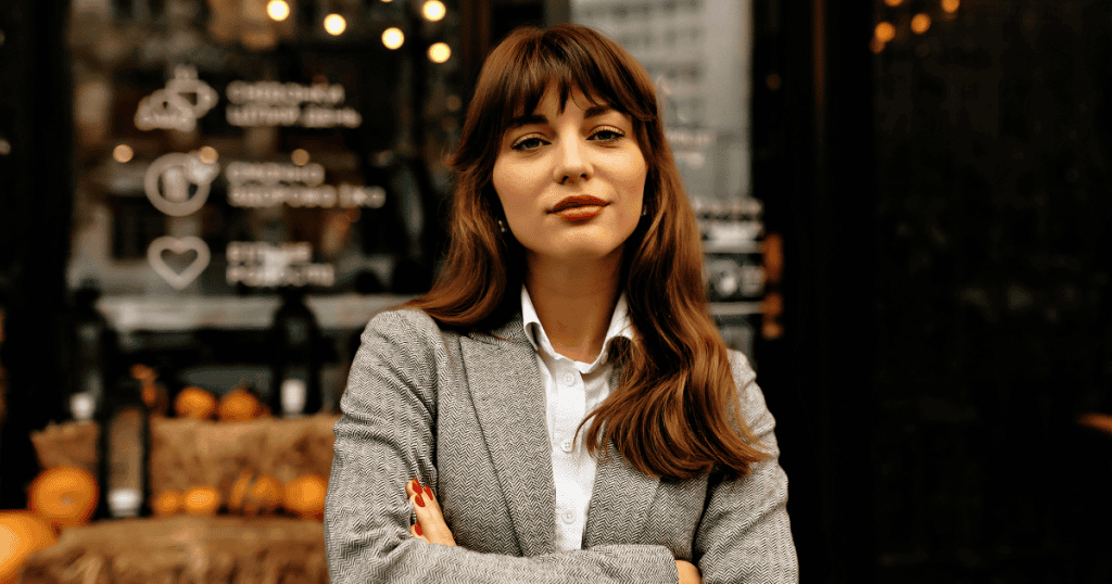 A woman with long brown hair and a blazer stands outdoors, arms crossed with a serious expression; her determined gaze suggests a powerful method for reversing the emotional cost.