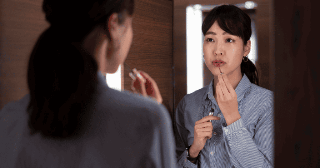 A woman in a blue pinstripe collared shirt applies lipstick while looking intently at her reflection in a mirror.