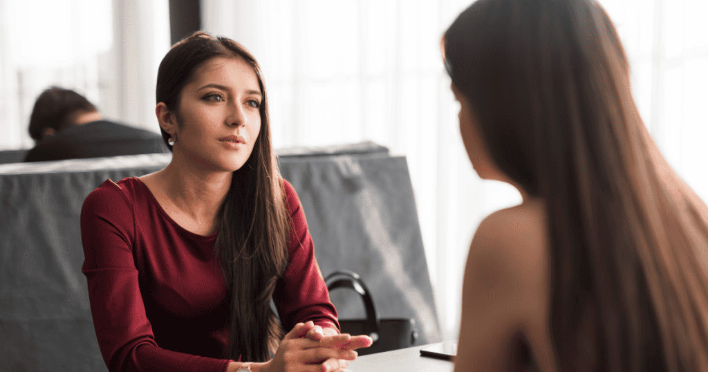A woman in a dark red top leans forward with an intense, focused expression while talking to someone off-camera; her gaze suggests she is seeking a deep, honest answer.
