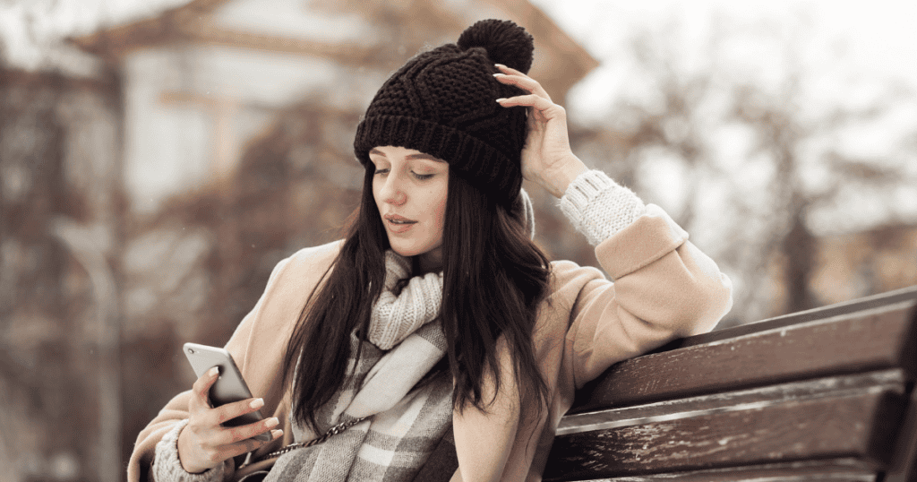 A young woman wearing a black knit hat and a tan coat sits on a park bench in a wintry setting, looking at her smartphone.