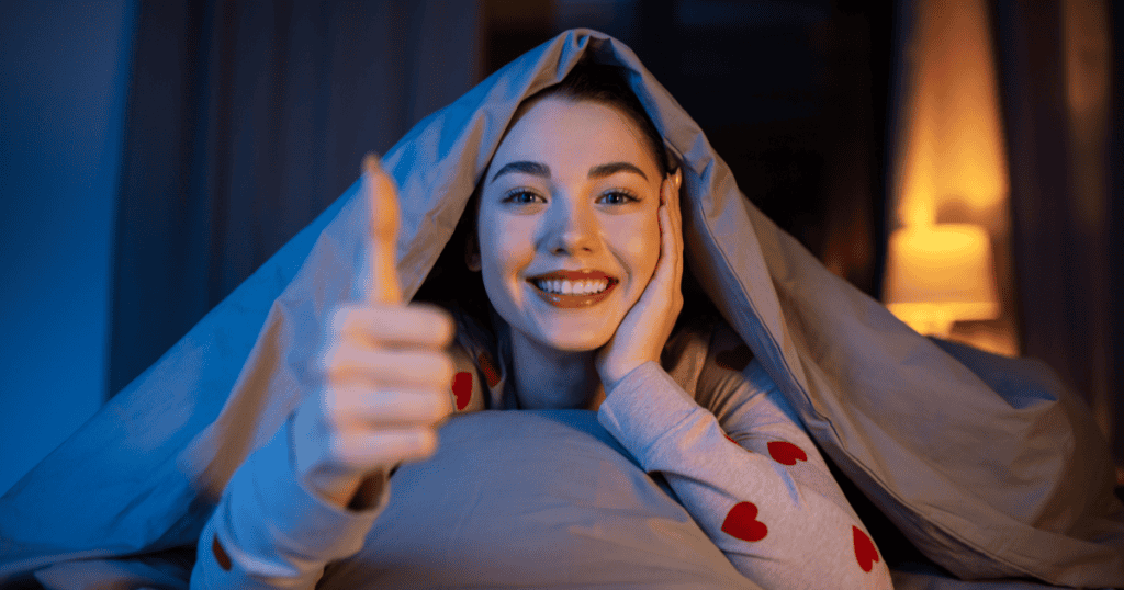 A smiling woman lying under a blanket gives a thumbs up, reflecting the habit of ending the day on a positive note to strengthen and protect emotional well-being.