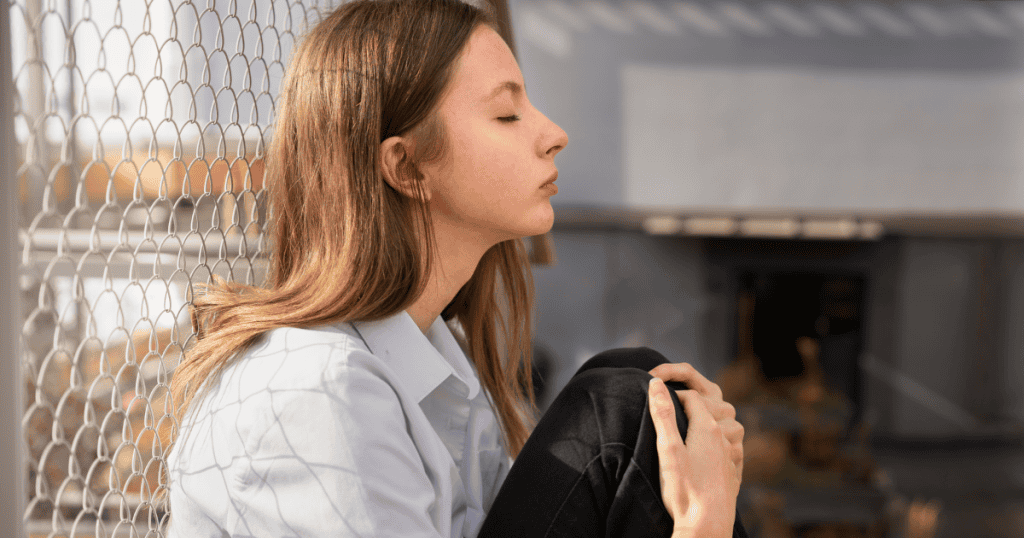 A young woman sits outdoors with her eyes closed and knees pulled close, symbolizing guilt as the emotional trap that keeps you small.