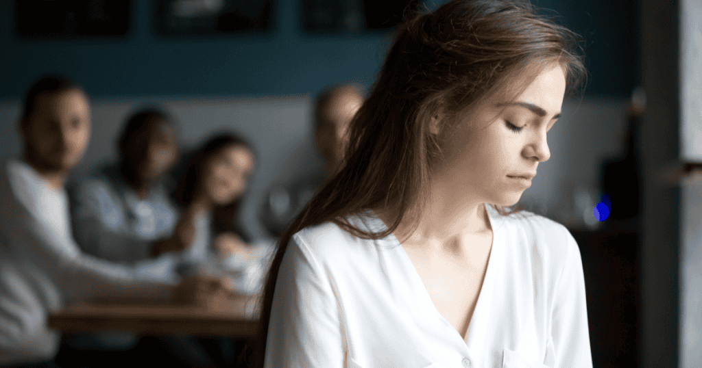 A young woman sits apart, eyes closed in pain, while others in the background watch, reflecting the weight of unhealthy guilt imposed by narcissists.