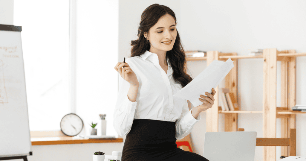 A smiling woman in a white blouse and black skirt sits and holds a pen and papers; her careful, contemplative look is key to executing powerful maneuvers without risk.