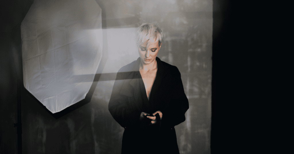 A woman with short blonde hair looks down at her phone in a dimly lit studio with a softbox, showing a moment of focused, internal resolve to maintain one's guard.