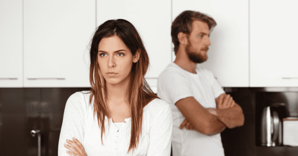 A woman stands confidently with her arms crossed while a man sulks in the background, capturing the quiet strength of someone realizing that a relationship built on constant agreement isn’t truly healthy.