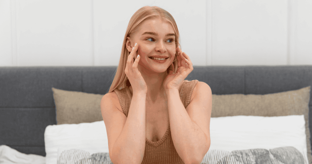 A smiling young woman with blonde hair, wearing a beige top, sits on a bed and gently touches her cheek.