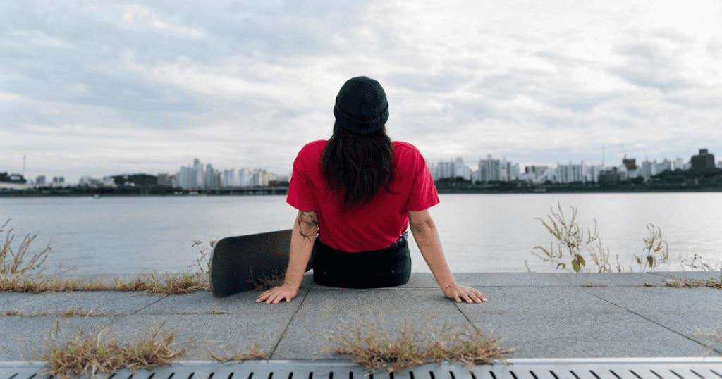 A young person wearing a red shirt and a dark knit cap sits on the edge of a waterfront, looking out at a city skyline across the water. A skateboard rests next to them.