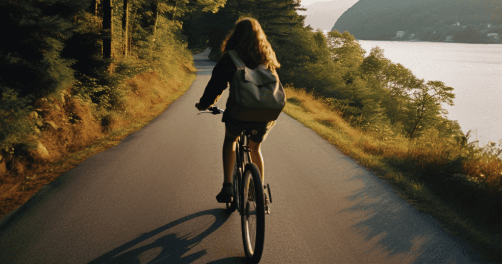 A woman with a backpack rides a bicycle on a scenic road beside a lake and trees, illustrating the idea to move like your brain depends on it.