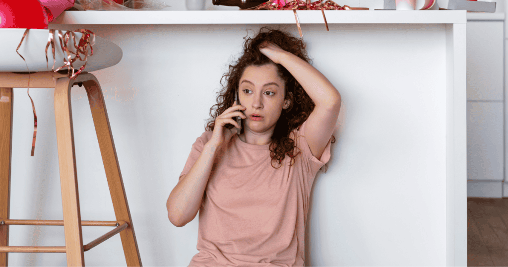 A young woman with curly hair sits on the floor amidst party remnants, looking stressed while talking on a phone; she is trying to stabilize her situation despite the chaotic aftermath.