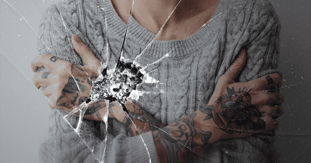 A tattooed person in a gray sweater crosses their arms tightly behind cracked glass, symbolizing obligation turning kindness into chains.