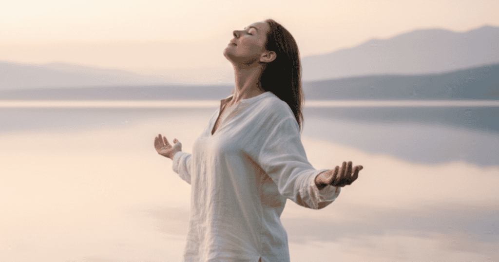 A woman stands outdoors with her eyes closed and arms open wide to a lake and sunset, embodying the liberating feeling that comes from releasing an excessive emotional load.