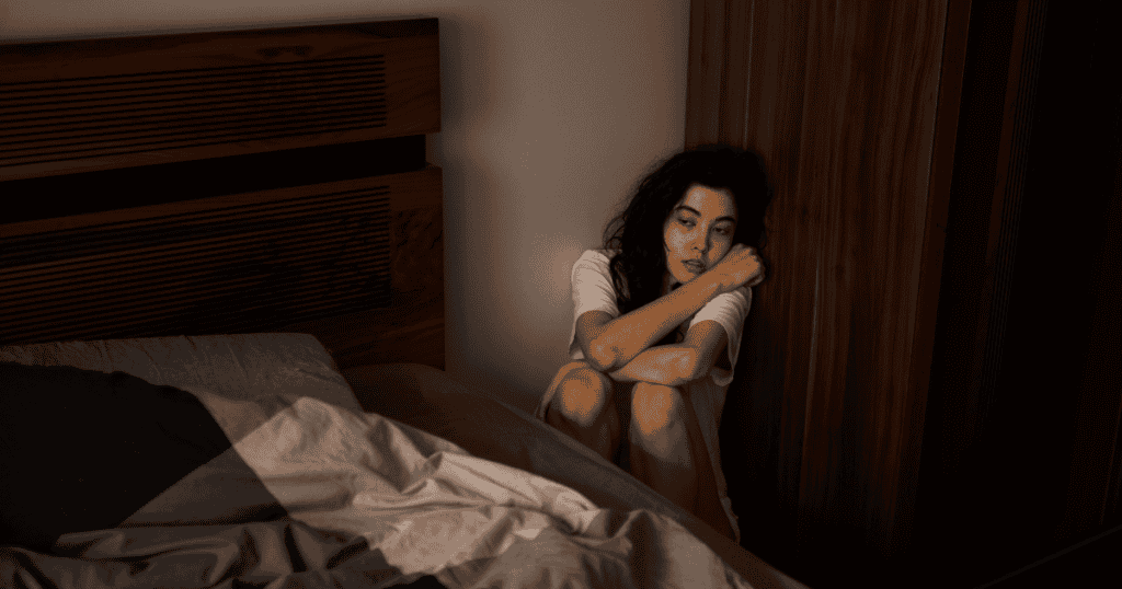 A woman sits on the floor beside her bed, looking lost in thought — a quiet reflection of the invisible, controlling rules survivors once had to live under.