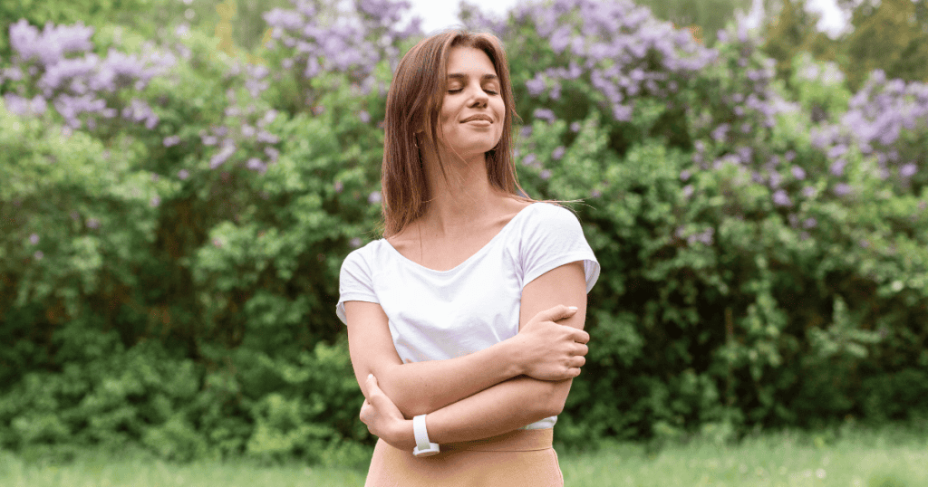 A smiling woman with her eyes closed stands in a park with her arms crossed, embracing herself; she has found a sense of security from within, rather than from external sources.