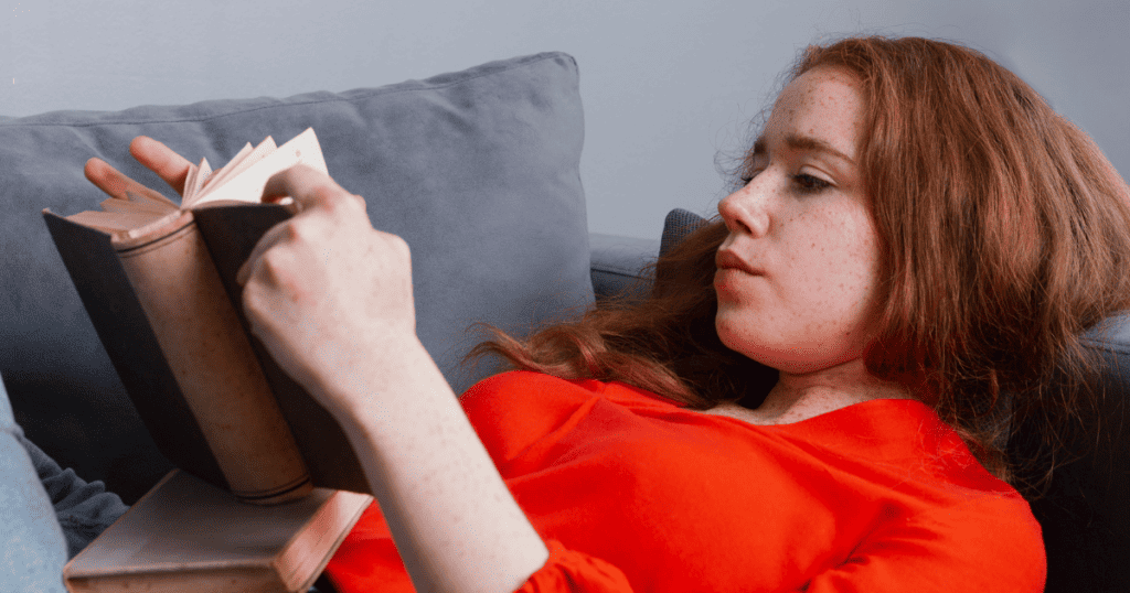 A young woman with red hair and freckles lies on a couch in a bright orange top, engrossed in reading a book; she is absorbing new narratives that challenge old, painful beliefs.