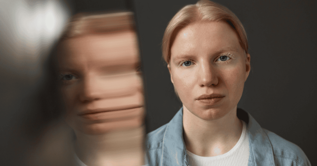 A woman with striking blonde hair and a denim jacket stares directly ahead, with a blurred, distorted image of her face next to her; she is challenging her automatic reactions to her inner feelings.