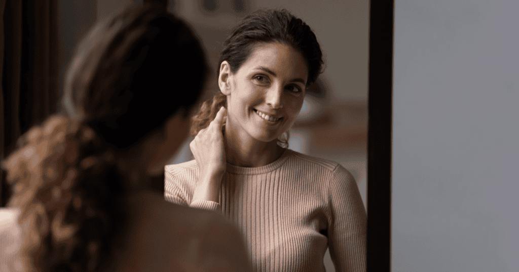 A woman smiles gently at her reflection in the mirror, radiating calm confidence and inner strength as she reconnects with her sense of self.