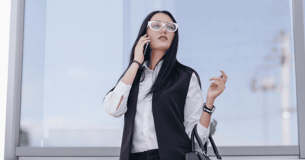 A woman in a white shirt and dark vest with white-rimmed glasses is speaking intensely on a cell phone outdoors, capturing a flash of highly reactive communication in an explosive moment.