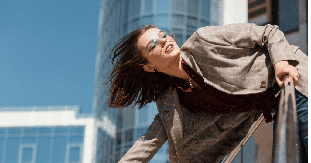 A woman in sunglasses and a plaid blazer leans over a railing in a cityscape, suggesting a high-rise view of planning and observation before action is taken.