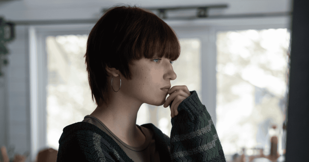 A close-up side profile of a person with short, dark red hair, wearing a green striped cardigan, looking thoughtful with their hand touching their chin.