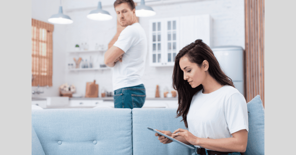 A woman focuses on her tablet while a man stands behind her with crossed arms, illustrating the subtle power shift when a narcissist begins exerting control.