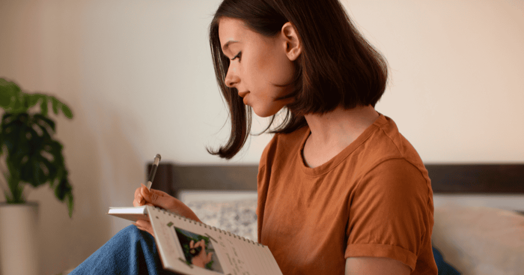 A young woman with short, dark hair sits on a bed writing in a notebook, deep in thought; this deliberate introspection is the first step toward self-discovery.