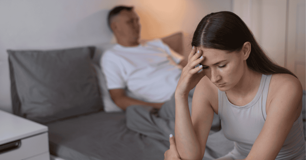A woman sitting on the edge of a bed looking distressed, while a man sits behind her, blurred in the background.