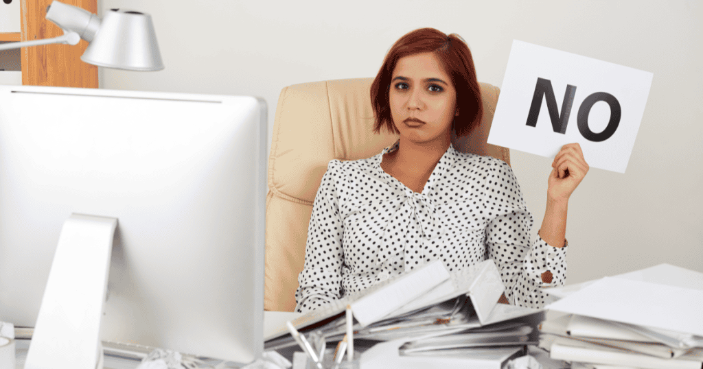 A woman sitting at her desk holding a paper sign that says โNO,โ showing rejection of the illusion narcissists create about their supposedly perfect new lives.