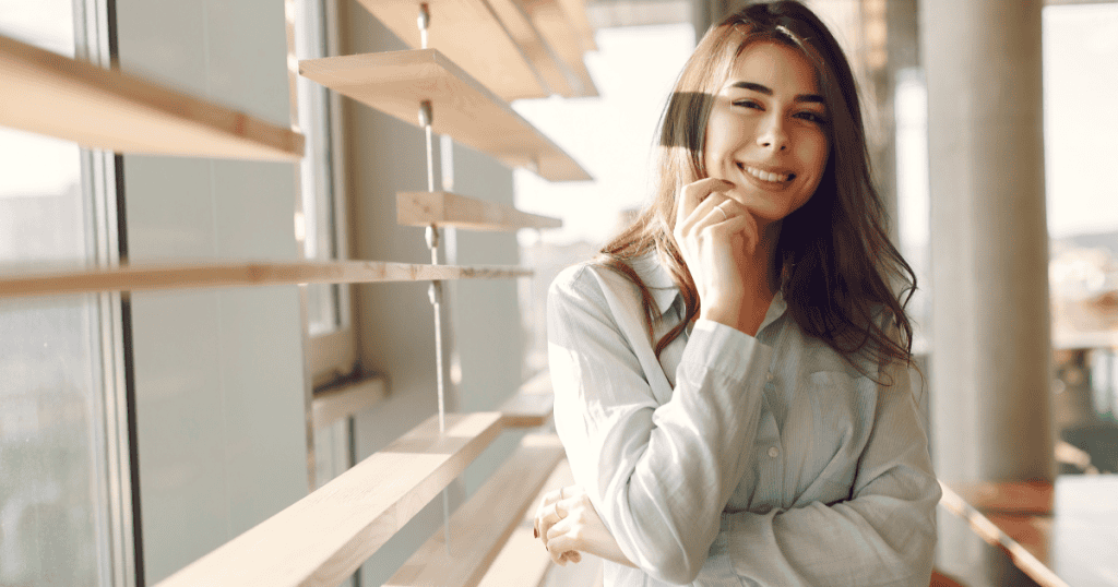 A confident woman stands by a sunlit window, smiling calmly and exuding self-assurance and healthy boundaries.