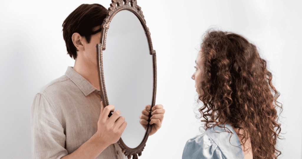 A man holds a mirror in front of his face while a woman looks at him, illustrating the idea of reflecting a narcissist’s behavior back to them to expose their own actions.