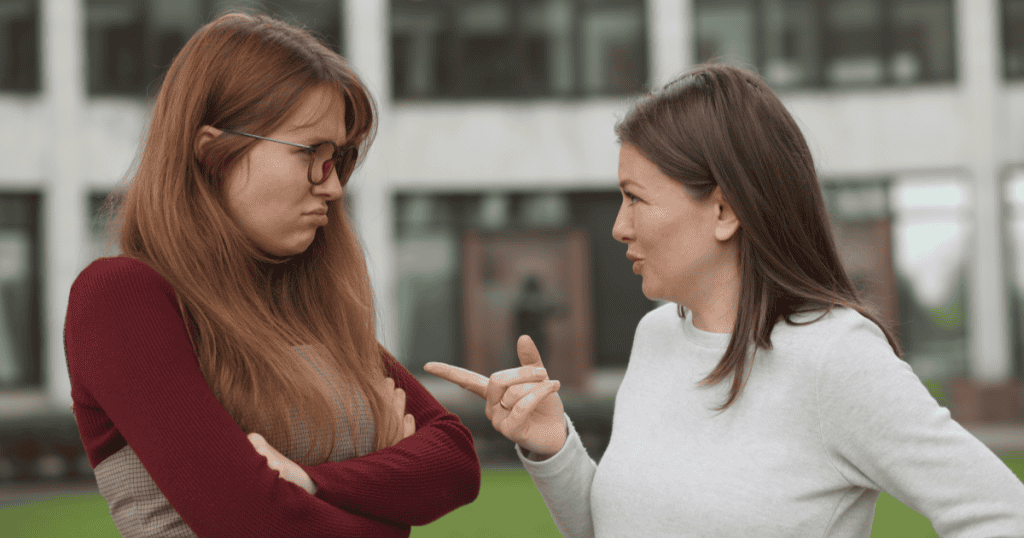 Two women stand facing each other in a tense conversation, one defensive with folded arms while the other questions her calmly, reflecting a power shift in a manipulative dynamic.