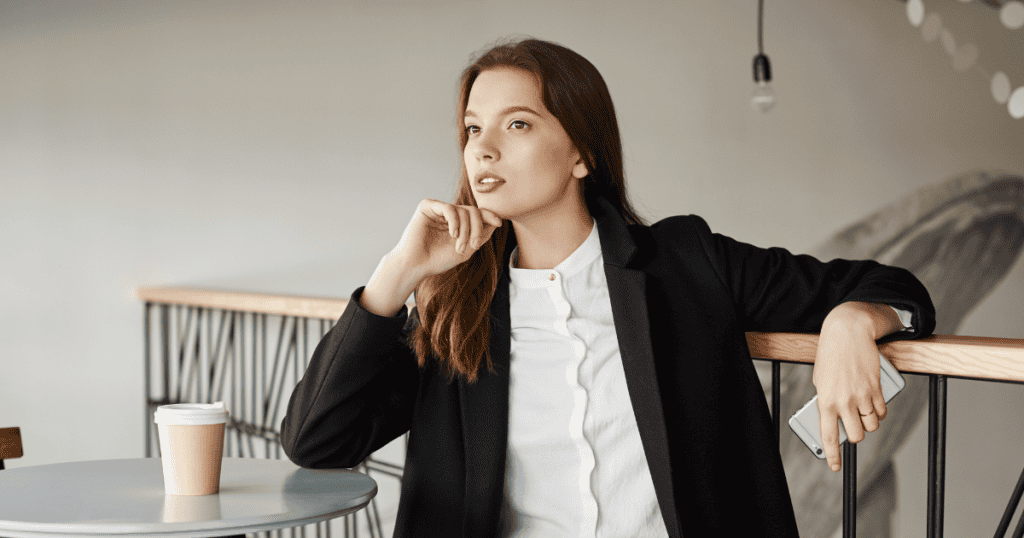 A confident woman in a blazer sits thoughtfully at a café table with a coffee cup and phone, representing the idea that your clarity is their downfall.