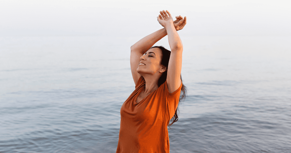 A woman stands by the water with her arms raised and a relaxed smile on her face, depicting a joyful return to a state of natural alignment and self-possession.