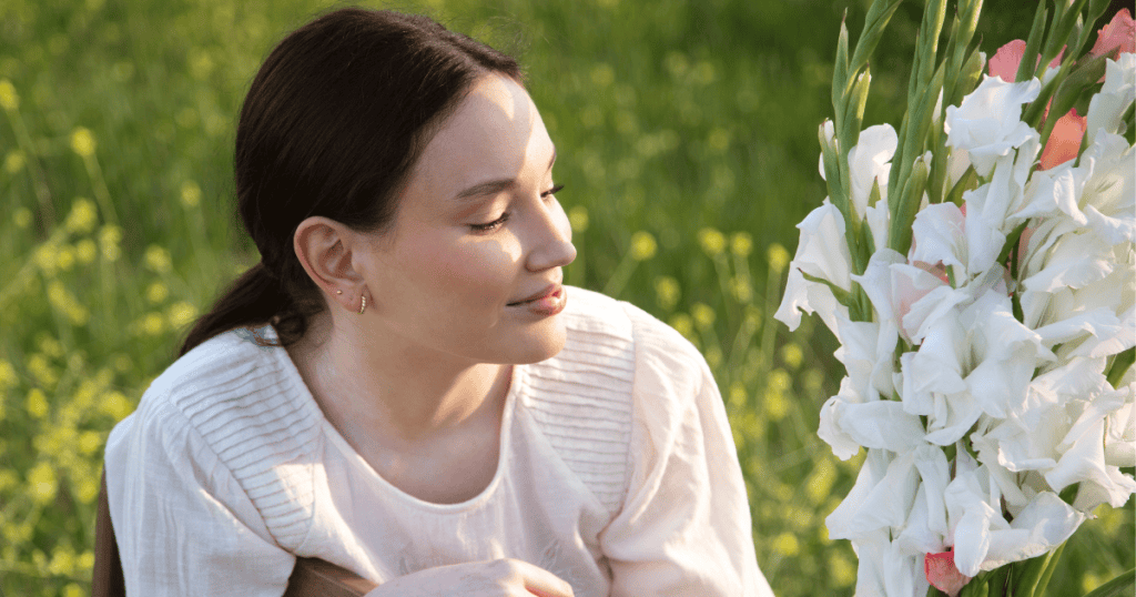 A serene young woman with light skin and a ponytail looks down at a bouquet of white flowers outdoors; she is connecting with her own peaceful, authentic self.